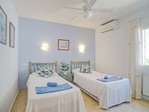 Two single beds with white bedspreads and blue towels, ceiling fan, air conditioning units, and a window with curtains.