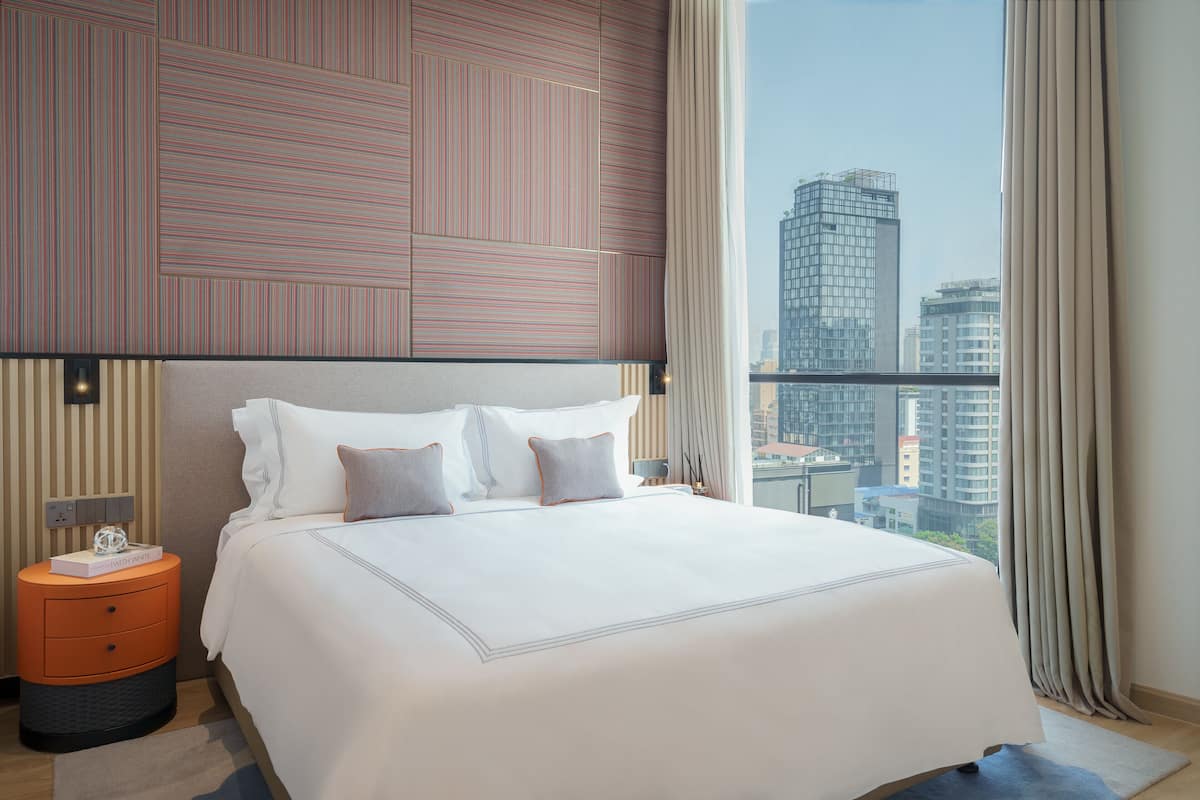 A modern hotel room with a large bed, a nightstand, a lamp, and a view of the cityscape through large windows.