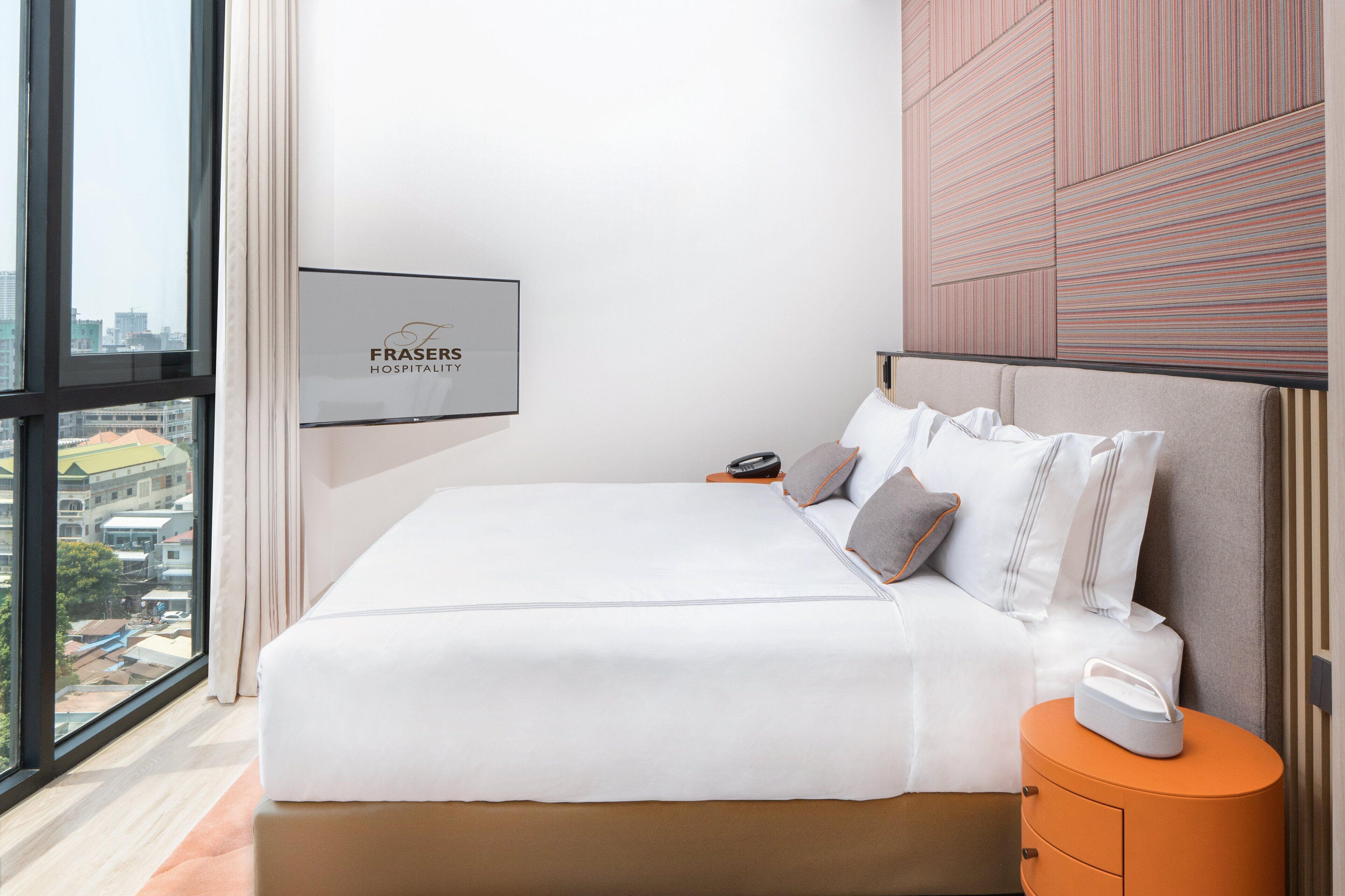 Una habitación de hotel moderna con una cama grande, una mesita de noche, un cartel enmarcado en la pared y vistas de la ciudad a través de amplios ventanales.