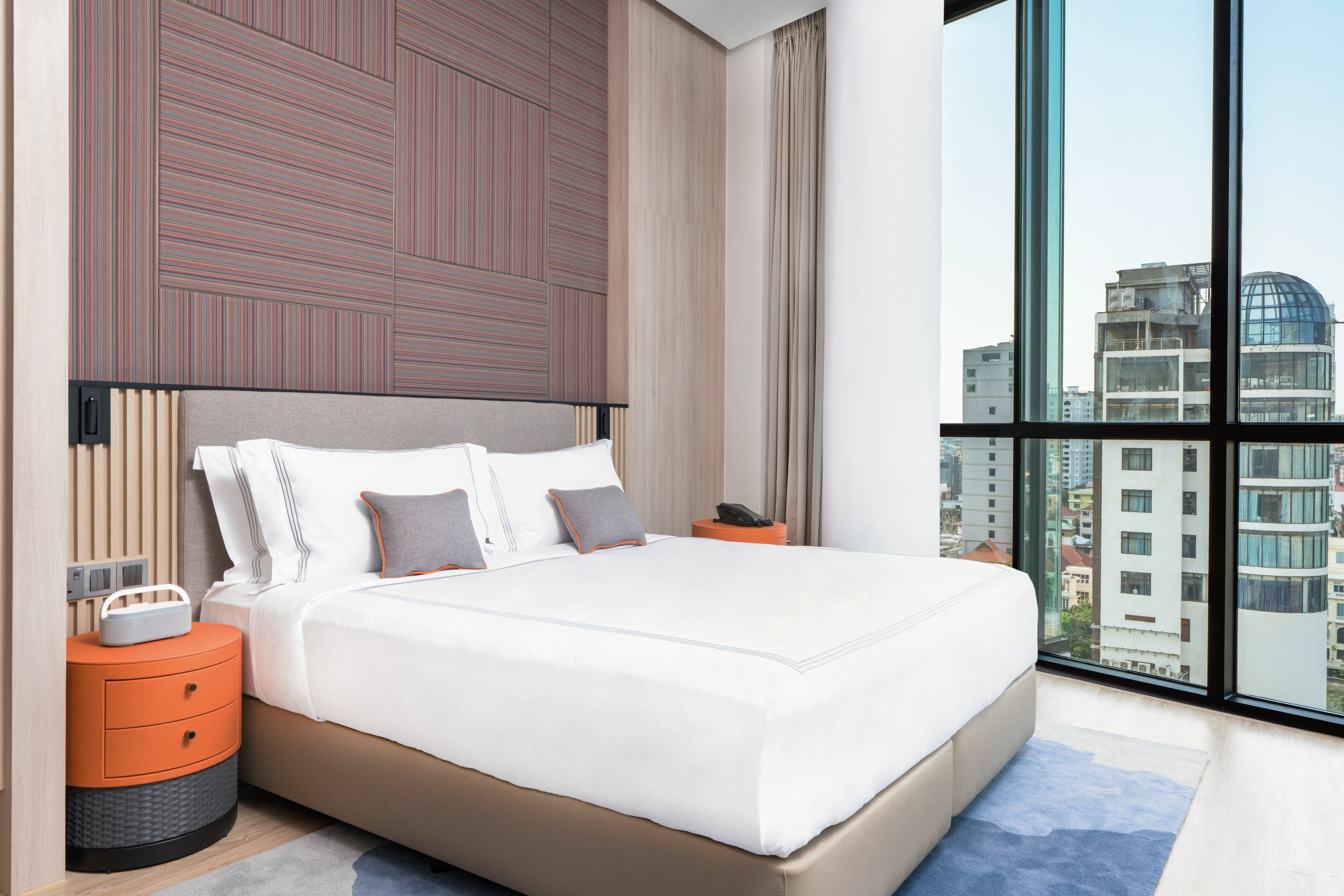 Ein modernes Hotelzimmer mit einem großen Bett, einem Nachttisch und Blick auf die Stadt durch große Fenster.