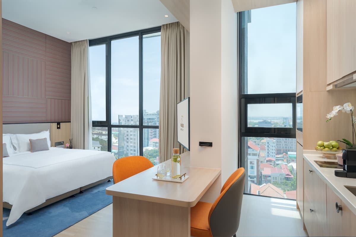 A hotel room with a large bed, a desk with a chair, a view of the city through large windows, and a kitchen area with a sink and cabinets.