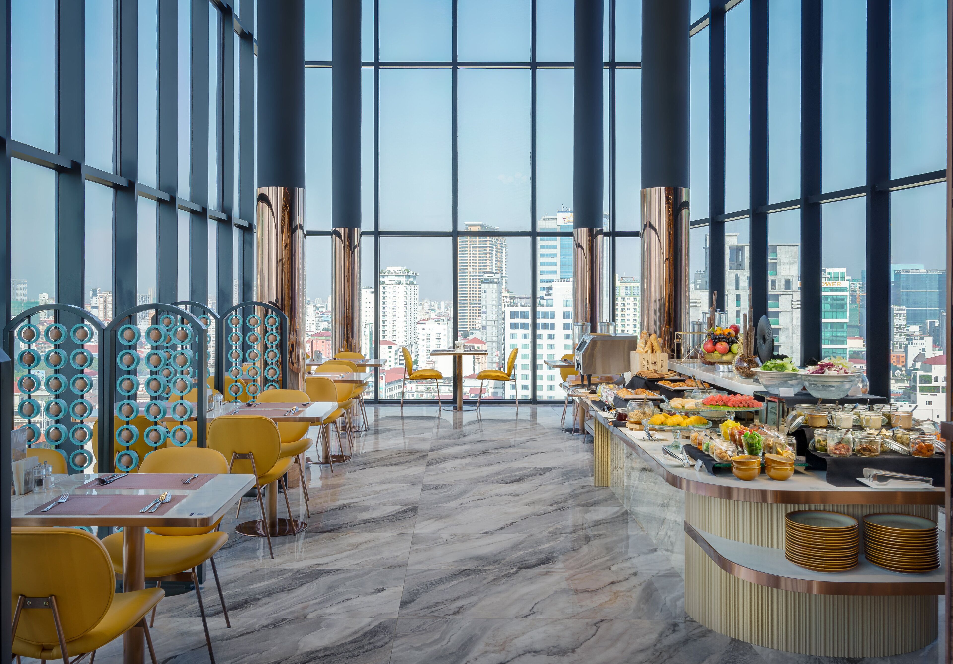 A modern restaurant with yellow chairs and tables, a buffet with various food items, and a city view through large windows.