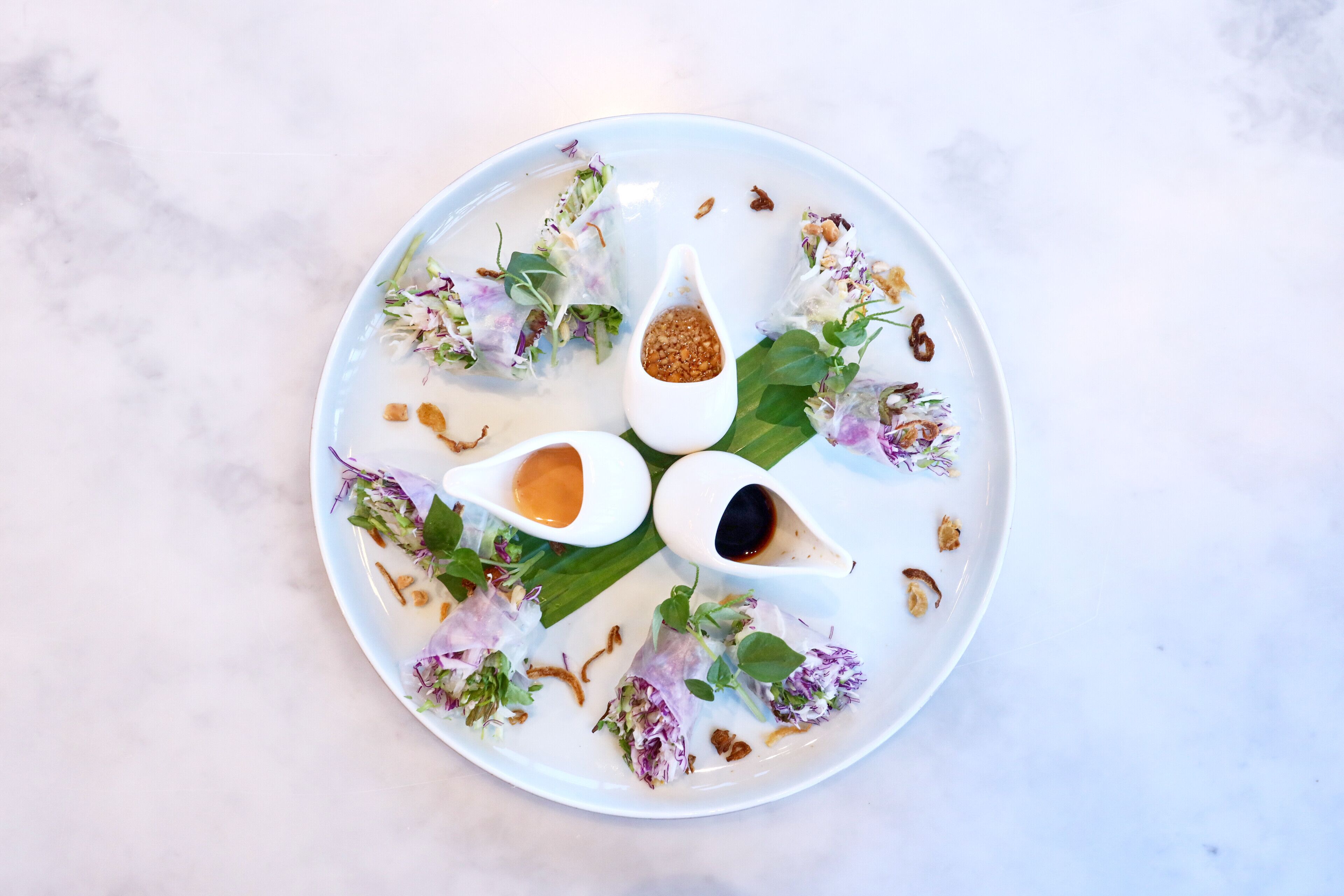 A plate with spring rolls, two dipping sauces, and garnishes on a marble surface.