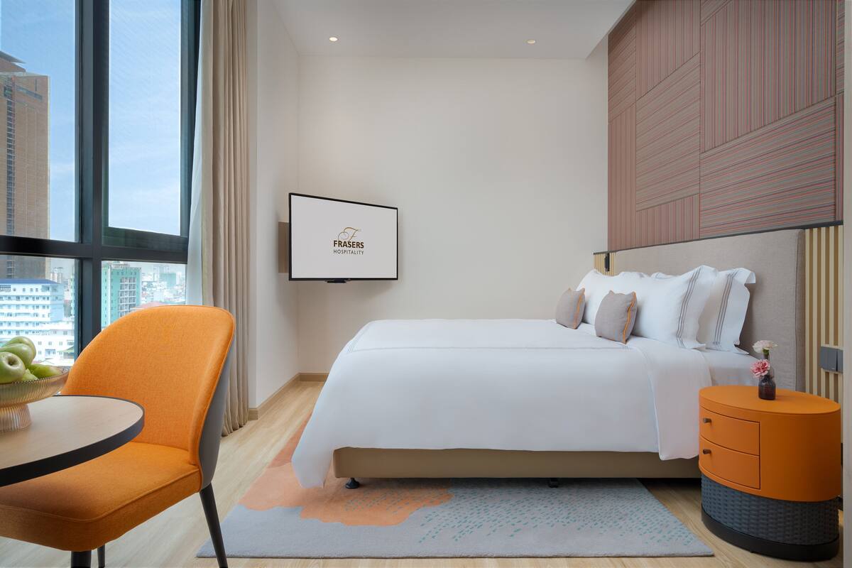 A hotel room with a bed, a TV mounted on the wall, a round orange table, and a chair.