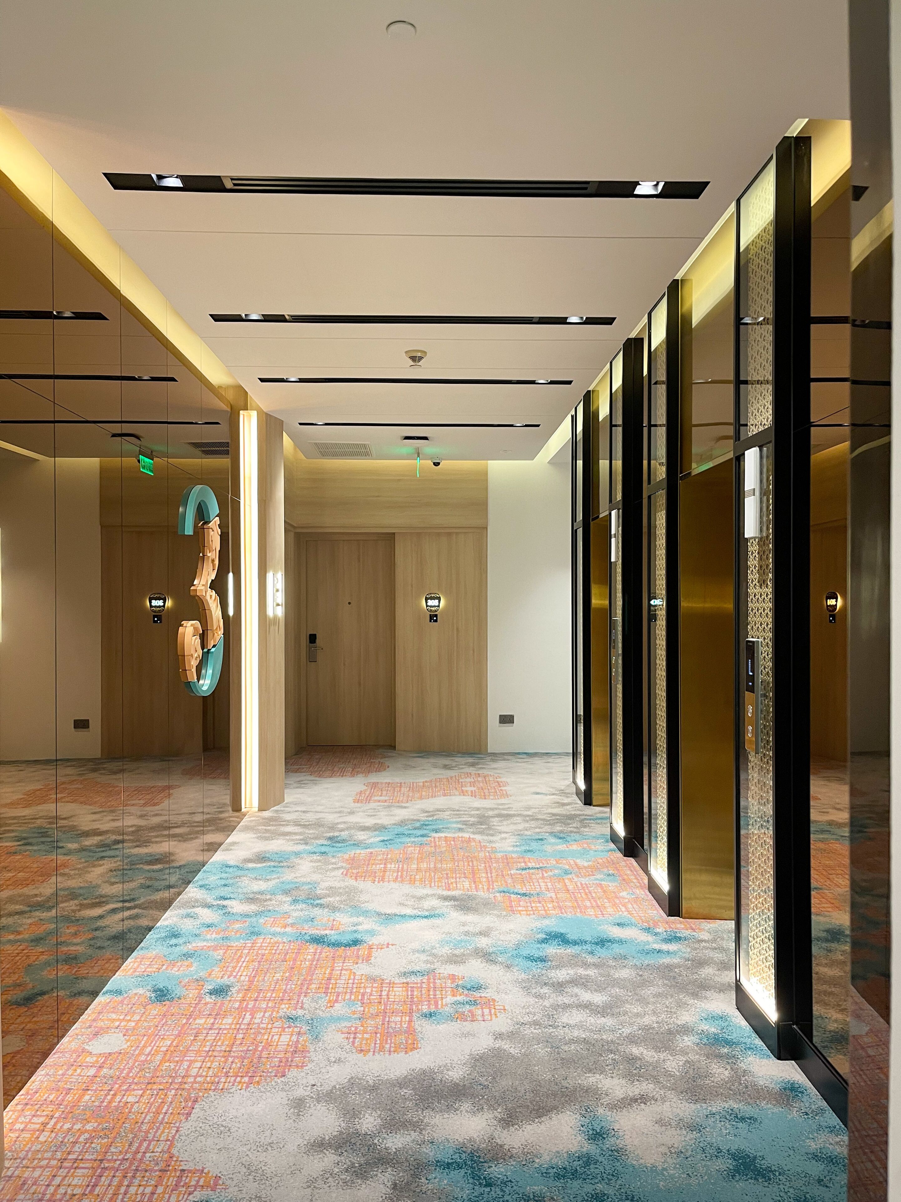 A modern hotel lobby with a patterned carpet, wooden panel walls, and a glass elevator.