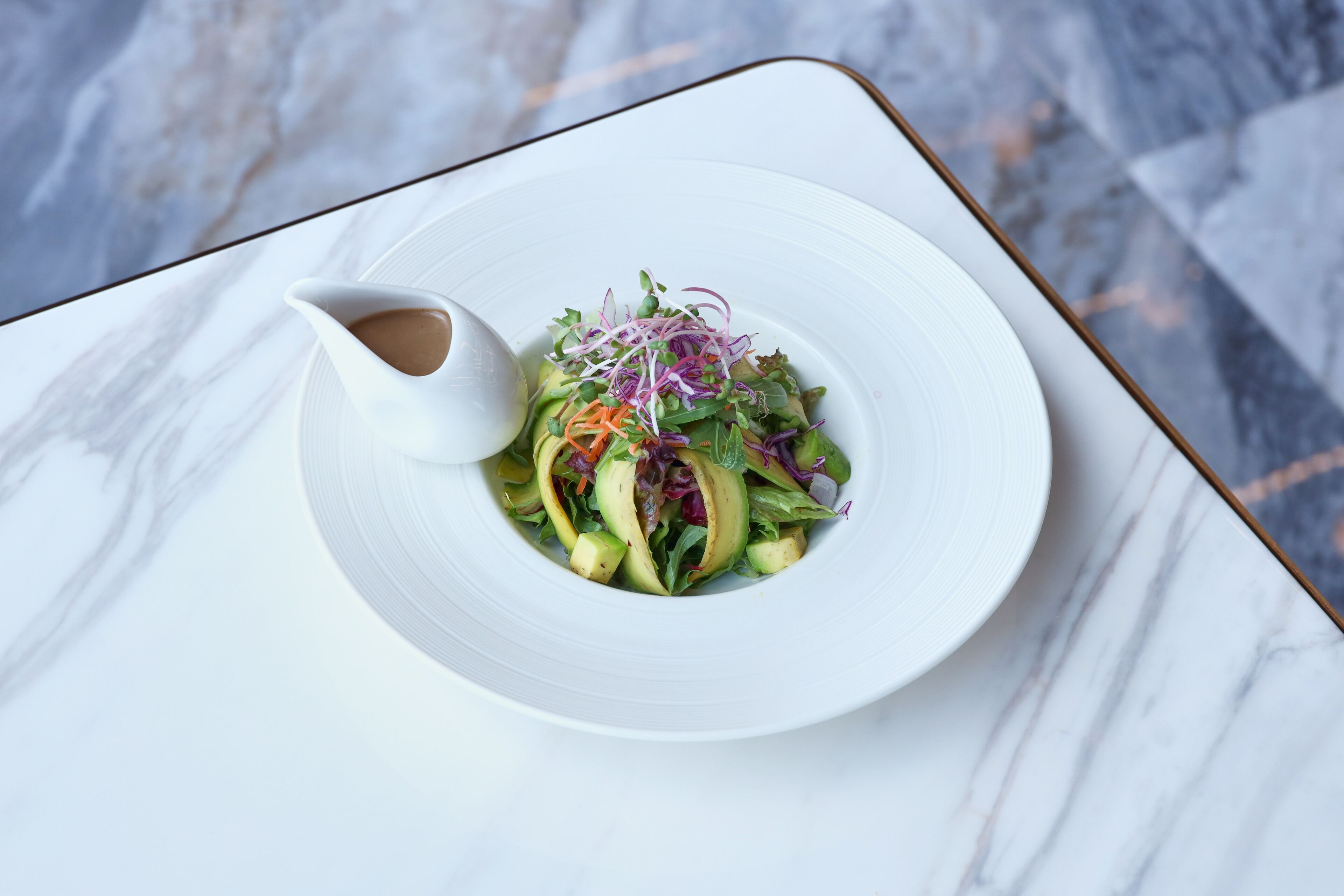 A white plate with a salad and a small white bowl of dressing on a marble table.