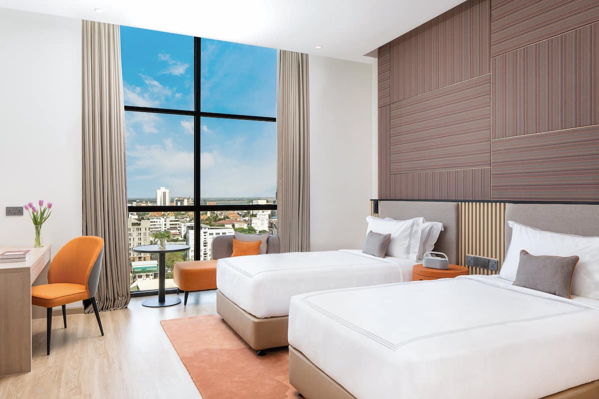 A hotel room with two beds, a desk, a chair, a small table, and a view of the city.