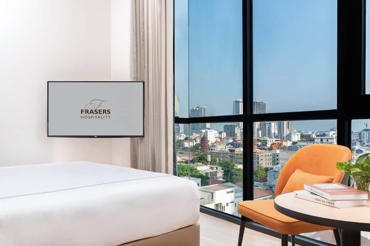 A hotel room with a bed, a chair, a table with books, and a framed sign on the wall.