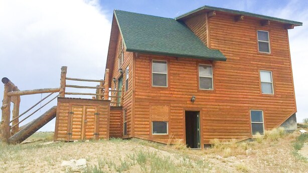 A two-story log cabin with a green roof, a wooden porch, and a staircase leading to the entrance.
