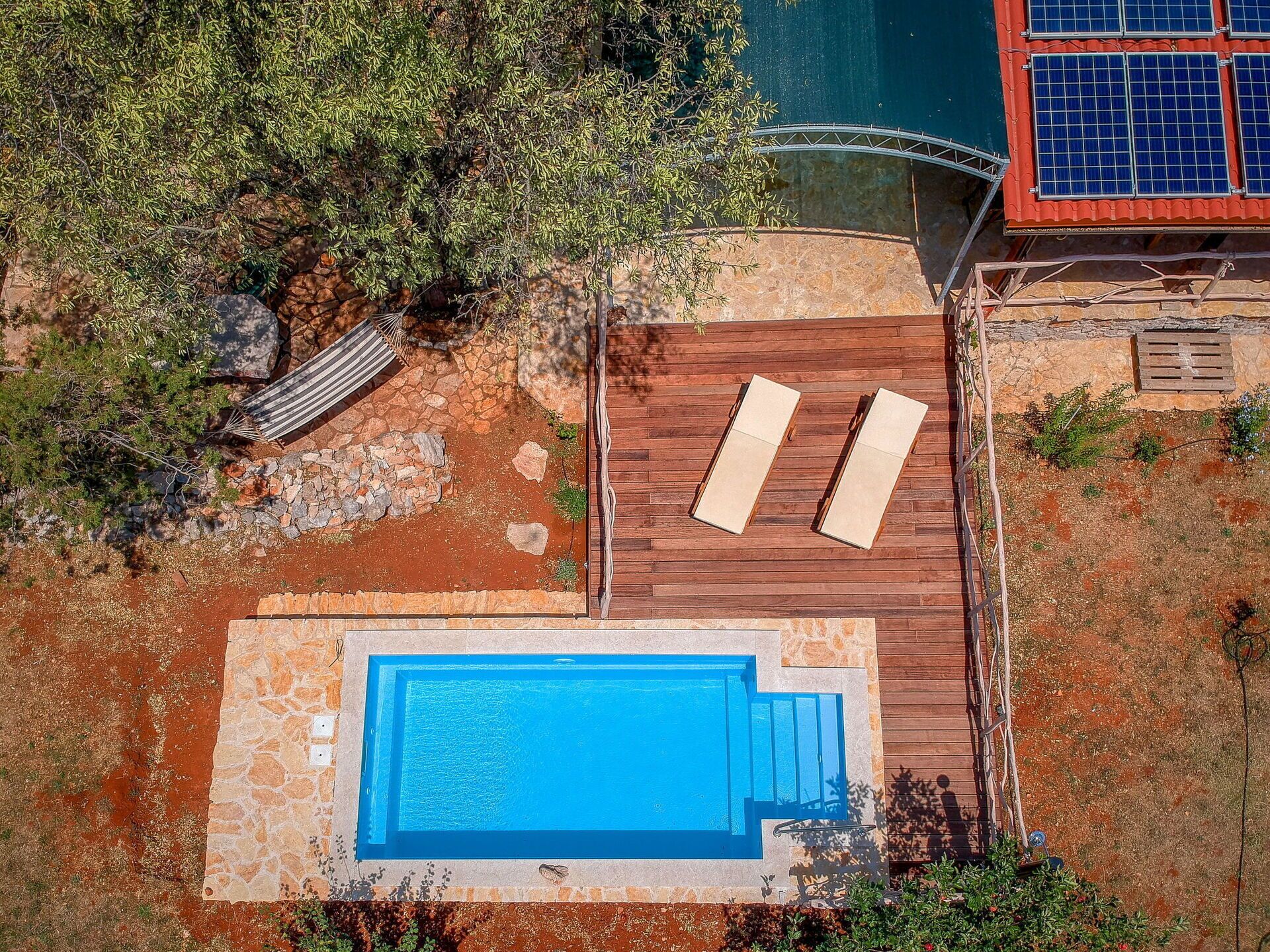 An aerial view of a swimming pool with wooden deck and lounge chairs, surrounded by greenery and a stone wall.