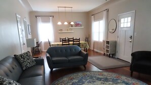 A living room with a blue sofa, patterned pillows, a round rug, a dining area with a table and chairs, and a kitchen area with a hanging light fixture.