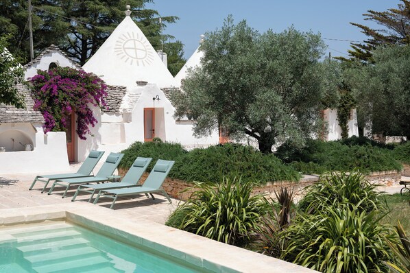 Un área de piscina con sillas de descanso, un edificio blanco con un símbolo de sol y abundante vegetación circundante.