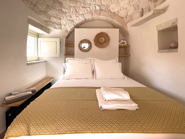 Un dormitorio con cama, almohadas, una toalla doblada, una mesita de noche de madera, un espejo y un estante con objetos decorativos.