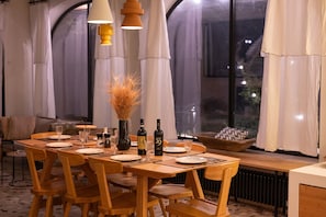 Ruokailutila, jossa on puinen pöytä, johon on katettu neljälle, kaksi viinipulloa, maljakko kuivine kasveineen ja näkymä ulos suurista ikkunoista.