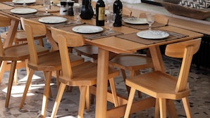 A dining area with a wooden table set for six, surrounded by wooden chairs, with a vase of dried plants and a bottle of wine on the table.