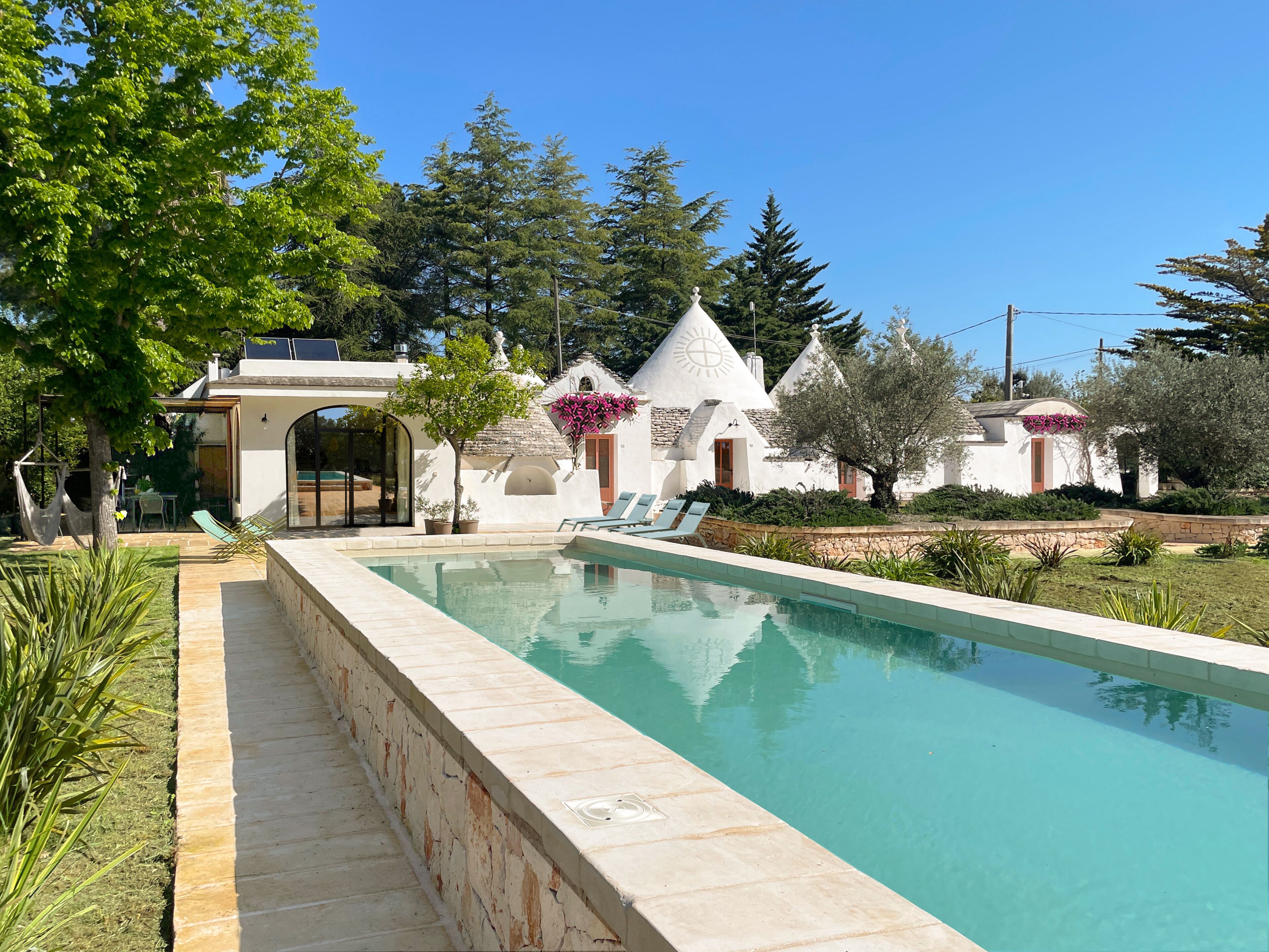 Una piscina con borde de piedra, rodeada de vegetación y edificios blancos con techos cónicos.
