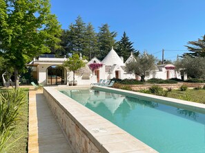 A swimming pool with a stone border, surrounded by greenery and white buildings with conical roofs. - Trullo Panna Fragola - Private Villa with 4 Double Rooms and a Swimming Pool (Martina Franca)