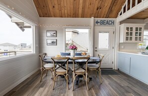 Uma área de refeições com teto de madeira, mesa com cadeiras, porta com a inscrição "BEACH" e vista para o exterior.