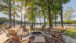 Een houten terras met een ronde vuurkuil en zitgedeelte, omgeven door bomen, met uitzicht op een meer.