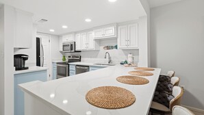 A modern kitchen with white cabinetry, a central island, and a dining area with chairs. - Beach Front, Balcony access from den and master, Spacious living, fully equipped (Gulf Shores)