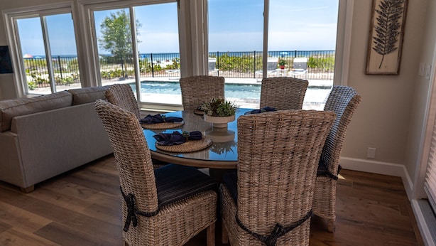 A dining area with wicker chairs and a glass table, overlooking a pool and a beach. - Direct Oceanfront Oasis: Remodeled Home with Private Heated Pool (Indian Shores)