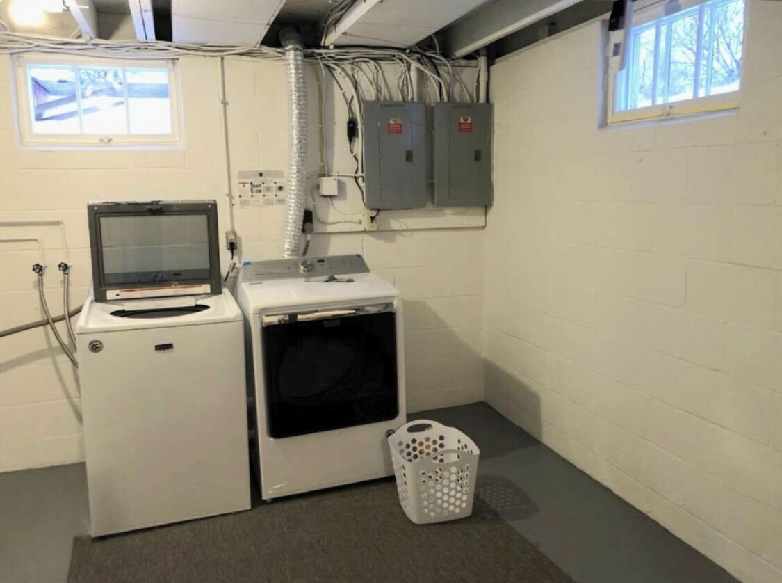 Une buanderie équipée d’un lave-linge, d’un sèche-linge et d’un petit écran. On y trouve des boîtiers électriques fixés au mur et une fenêtre avec des stores.