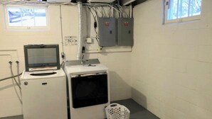 Un lavadero con lavadora, secadora y una pequeña pantalla. Hay cajas eléctricas en la pared y una ventana con persianas.