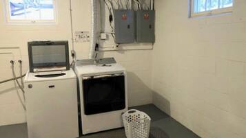 Une buanderie équipée d’un lave-linge, d’un sèche-linge et d’un petit écran. On y trouve des boîtiers électriques fixés au mur et une fenêtre avec des stores.