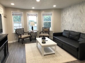 Un salon avec un canapé en cuir foncé, une table basse blanche, un fauteuil à rayures et une petite table ronde avec une lampe. Il y a trois fenêtres équipées de stores, un téléviseur fixé au mur et un meuble en bois.