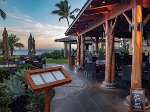 An outdoor seating area with wooden pillars, tables, and chairs, overlooking the ocean.