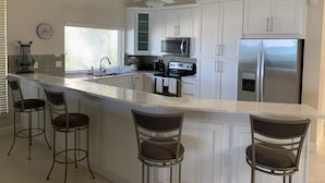 Una cucina moderna con mobili bianchi, una grande isola con sgabelli da bar, un forno a microonde, un frigorifero e una ventola a soffitto.