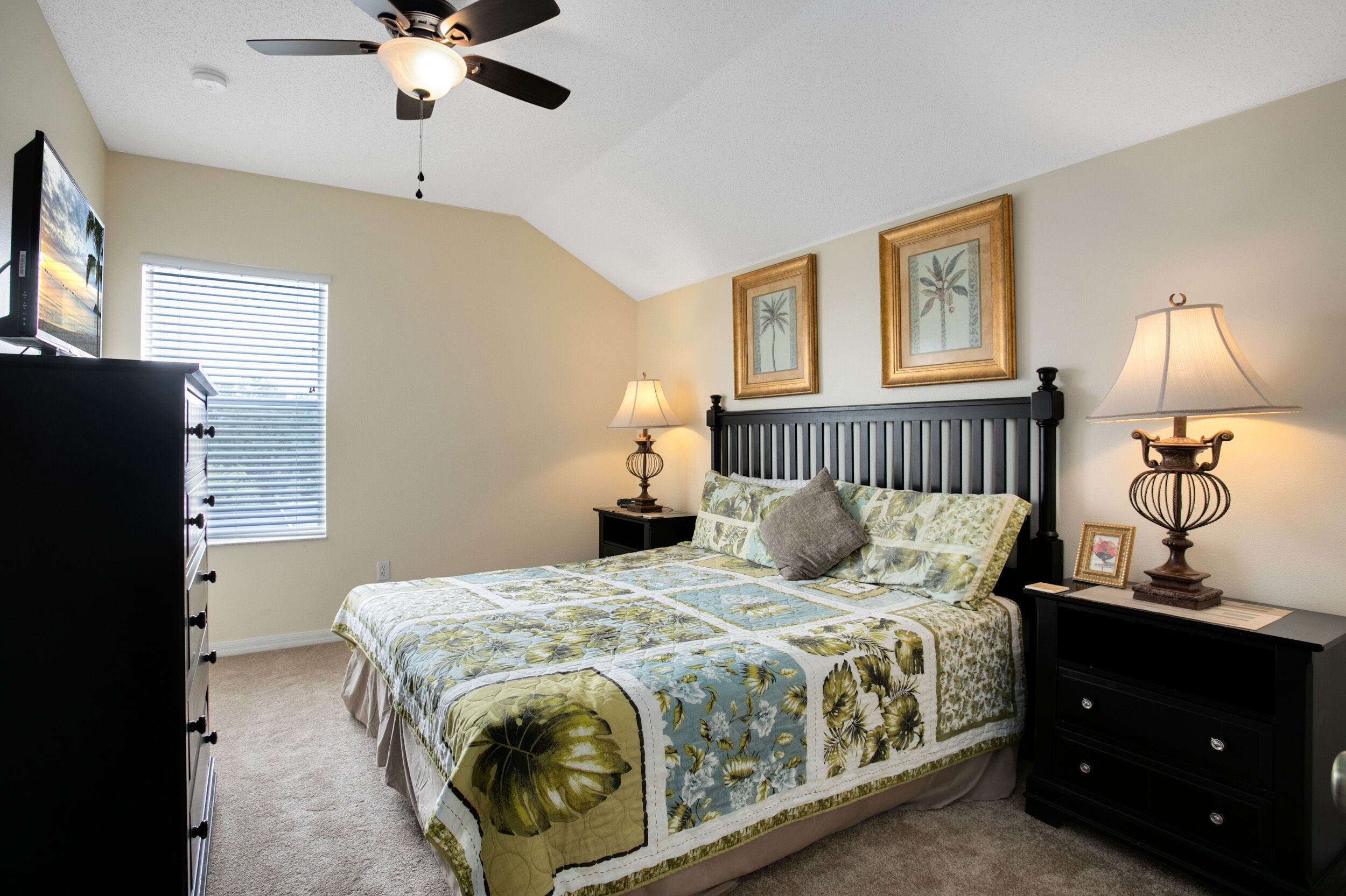 A bedroom with a bed, a ceiling fan, a nightstand with a lamp, and a dresser.