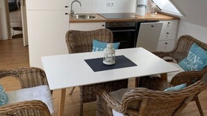 Una cocina compacta con encimera blanca, un lavamanos y una cafetera. Hay una mesa de comedor con sillas de mimbre y un farol sobre ella.