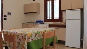 Een keuken met een tafel bedekt met een tafelkleed met een fruitprint, houten stoelen, een witte koelkast en een raam met houten luiken.