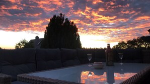 Un área de estar al aire libre con una mesa de vidrio, cojines y una botella de vino, con el cielo al atardecer de fondo.
