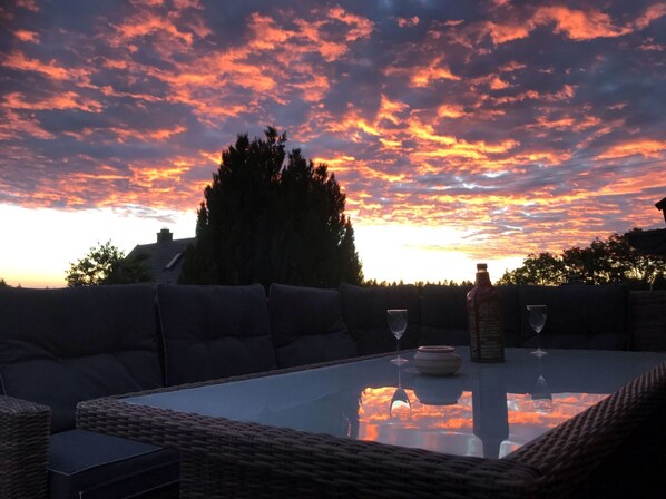 An outdoor seating area with a glass table, cushions, and a bottle of wine, set against a sunset sky.