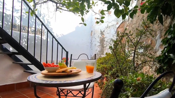 Een tafel met een schaal fruit, een broodplateau en een glas jus d'orange op een balkon met uitzicht op een rotsachtige klif en groen.
