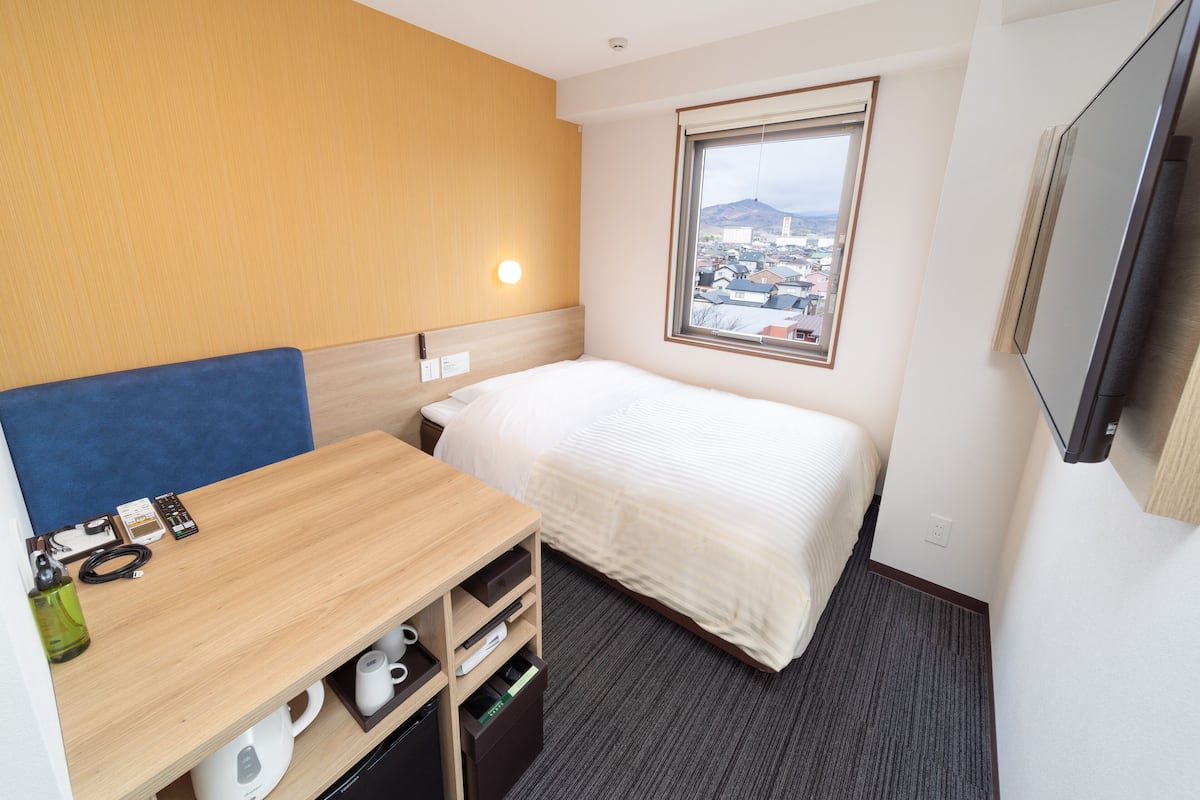 A hotel room with a bed, a desk with a chair, a small table, a TV, and a window with a city view.