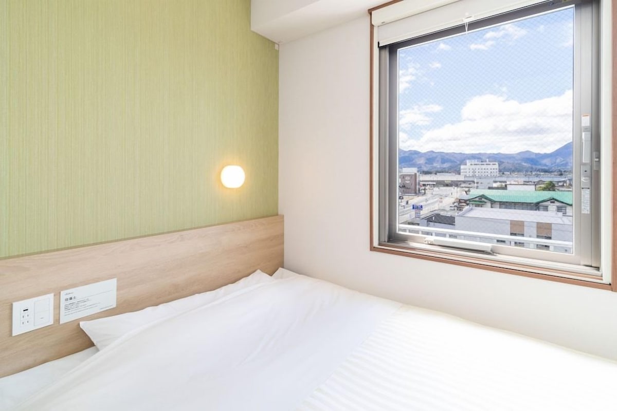 Habitación de hotel con cama, ventana que ofrece vistas a edificios y montañas, y una lámpara fijada a la pared.