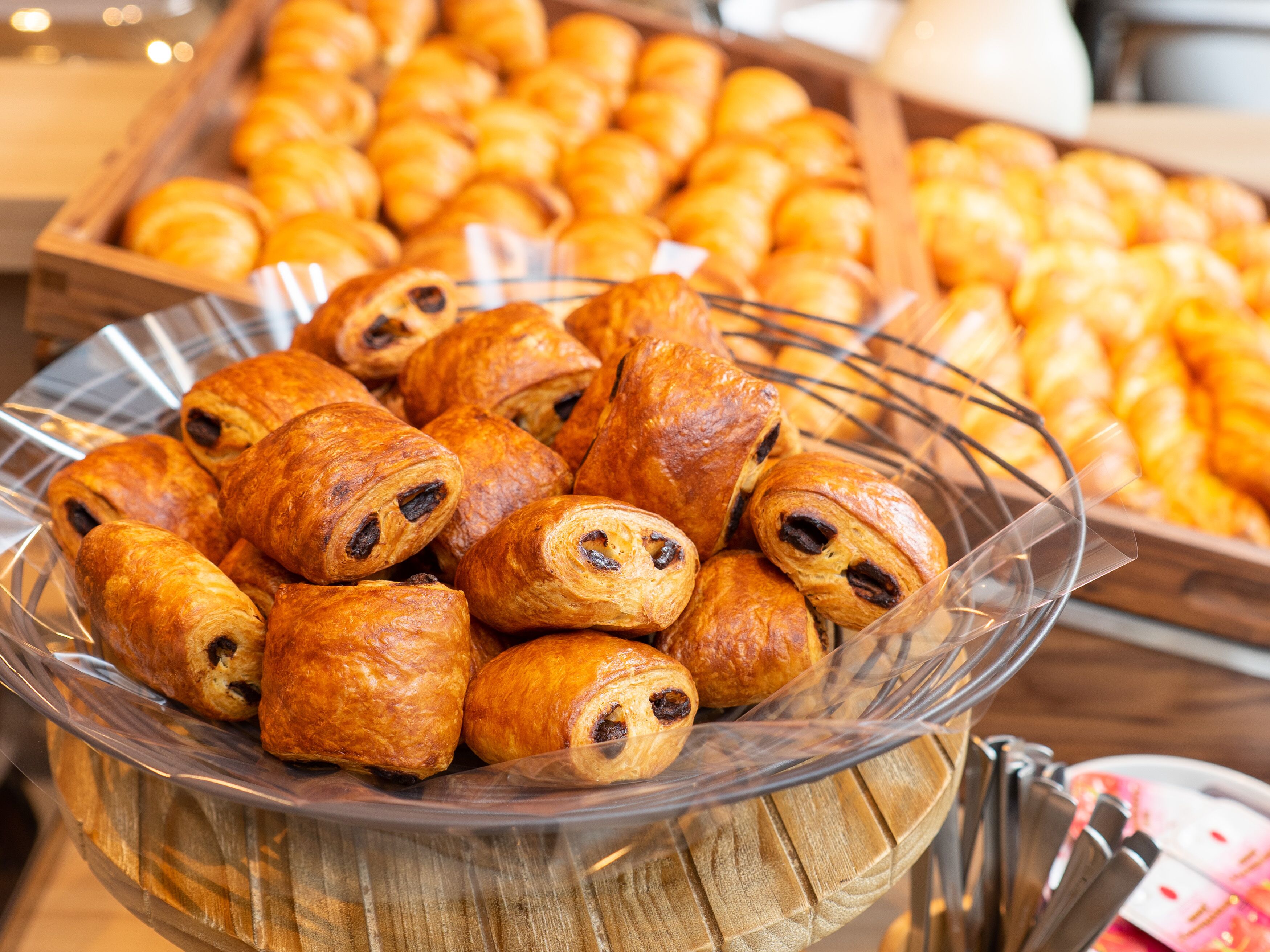 A glass bowl filled with assorted pastries, including croissants and chocolate-filled rolls, displayed on a wooden stand.