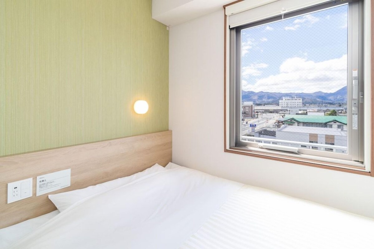 Habitación de hotel con cama, ventana que ofrece vistas a edificios y montañas, y una lámpara fijada a la pared.