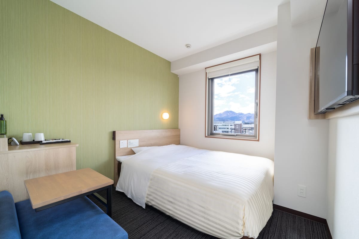 A hotel room with a bed, a desk with a telephone and cups, a blue sofa, a window with a mountain view, and a TV mounted on the wall.