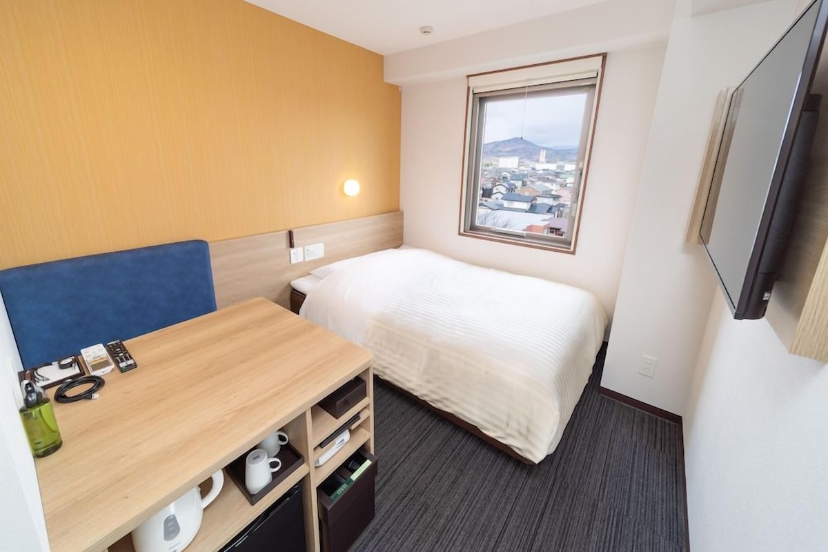 Habitación de hotel con una cama, un escritorio con silla, televisión y ventana con vista a la ciudad.