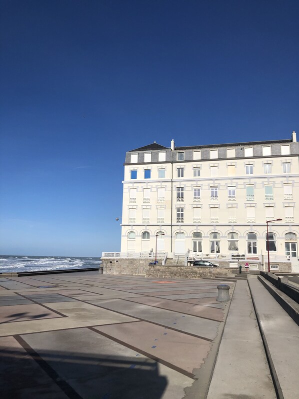 A large, multi-story building with a symmetrical facade, a prominent entrance with steps, and a view of the sea in the background.