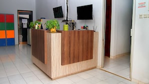 A reception desk with a wooden finish, two monitors, a plant, and a small table with items. - Hotel Syariah Mama Kutajaya Pasar Kemis (PasarKemis)