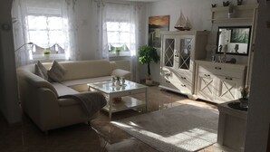 Un salón con sofá, una mesa de centro y un aparador. Hay ventanas con cortinas, una planta y un cuadro en la pared.