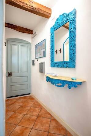 A hallway with a light blue door, a wall-mounted mirror with a blue ornate frame, a small shelf with a blue bowl, and a tiled floor. - Alguerhome CASA TURCHESE: a turquoise nest in the old town (Alghero)