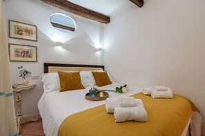 Una cama bien hecha con ropa de cama blanca y color mostaza, una mesita de noche con un florero y un libro, y un tragaluz en el techo.