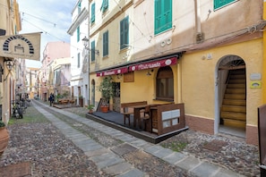 A narrow street with cobblestone pavement, a restaurant named 'Pizzeria,' and a building with a yellow facade and green shutters. - Alguerhome CASA TURCHESE: a turquoise nest in the old town (Alghero)