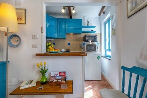 Cocina con armarios azules, un microondas y un refrigerador. Hay una ventana con contraventanas azules, un florero con flores y una mesa con un libro y una caja.